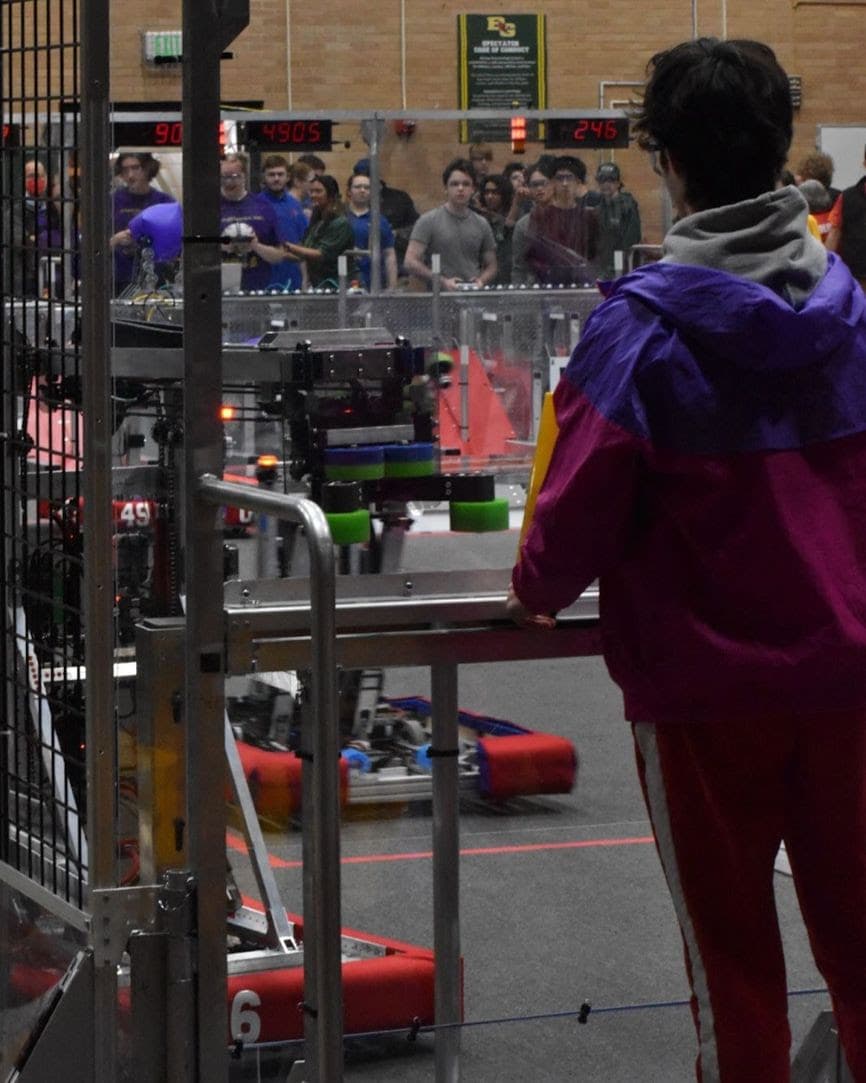 Owen '25 handing off a cone to our robot from the human player station during a match while George '24 and Maxwell '25 drive the robot in the background.